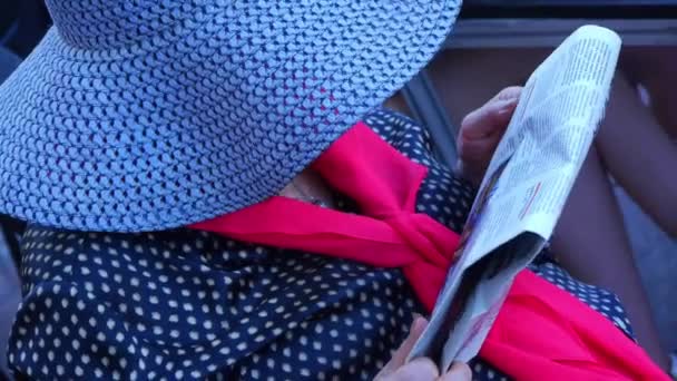 Een bejaarde vrouw in een hoed met brede rand en in een rode sjaal zit op een stoel en leest een krant — Stockvideo
