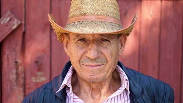 Retrato de un granjero anciano con sombrero de paja sobre un fondo de granja. La cara de un hombre de edad — Vídeo de stock