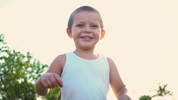 Un niño de 4 años con una camiseta blanca se ríe y extiende las manos. Retrato de un niño alegre y activo sobre un fondo natural . — Vídeo de stock