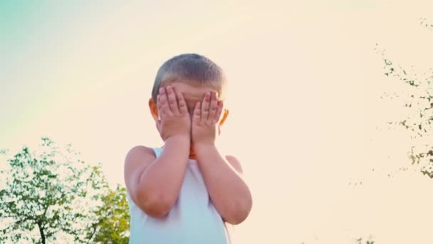 Rapaz de 4 anos com uma t-shirt branca ri, cobre o rosto com as mãos, o miúdo salta contra o céu. Retrato de uma criança ativa alegre em um fundo de natureza . — Vídeo de Stock