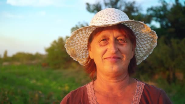 Πορτρέτο του μια ηλικιωμένη γυναίκα σε ένα καφέ μπλούζα και λευκό καπέλο σε φόντο τη καταπράσινη φύση το φως το βράδυ, η γυναίκα κοιτάζει την κάμερα και χαμόγελα, ο εργαζόμενος συγκομιδή για τη φυτεία — Αρχείο Βίντεο