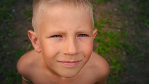 Retrato de cerca de un niño de ojos azules mirando y sonriendo directamente a la cámara, una mirada penetrante de un niño de 6 años, vista desde arriba — Vídeos de Stock