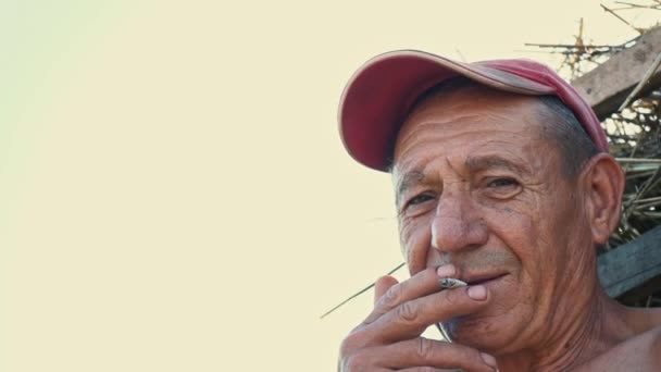 Porträt eines rauchenden Schwerarbeiters vor dem Hintergrund einer ländlichen Szene. Ein Bauer mit Mütze raucht eine Zigarette — Stockvideo