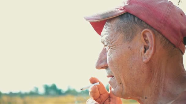 Portrait of a smoking hard worker against the background of a rural scene. A farmer in a cap smokes a cigarette — Stock Video