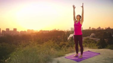 Genç bir kadın büyük bir şehir arka planda bir dağda yoga uygulamaları. Sağlıklı kadın günbatımında spor yapıyor. Kadın atlet pembe gömlek ve mor tayt güneş doğarken bir ısınma yok.
