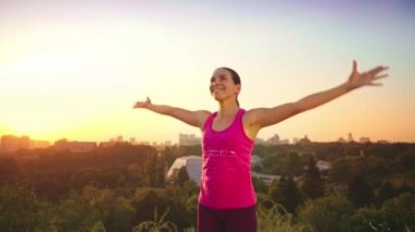 Genç bir kadın büyük bir şehir arka planda bir dağda yoga uygulamaları. Sağlıklı kadın günbatımında spor yapıyor. Kadın atlet pembe gömlek ve mor tayt güneş doğarken bir ısınma yok.