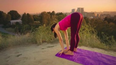 Genç bir kadın büyük bir şehir arka planda bir dağda yoga uygulamaları. Sağlıklı kadın günbatımında spor yapıyor. Kadın atlet pembe gömlek ve mor tayt güneş doğarken bir ısınma yok.