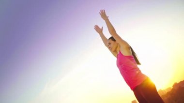 Genç bir kadın büyük bir şehir arka planda bir dağda yoga uygulamaları. Sağlıklı kadın günbatımında spor yapıyor. Kadın atlet pembe gömlek ve mor tayt güneş doğarken bir ısınma yok.
