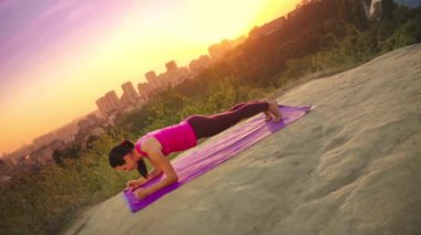 Genç bir kadın büyük bir şehir arka planda bir dağda yoga uygulamaları. Sağlıklı kadın günbatımında spor yapıyor. Kadın atlet pembe gömlek ve mor tayt güneş doğarken bir ısınma yok.