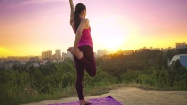Genç bir kadın büyük bir şehir arka planda bir dağda yoga uygulamaları. Sağlıklı kadın günbatımında spor yapıyor. Kadın atlet pembe gömlek ve mor tayt güneş doğarken bir ısınma yok.