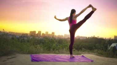 Genç bir kadın büyük bir şehir arka planda bir dağda yoga uygulamaları. Sağlıklı kadın günbatımında spor yapıyor. Kadın atlet pembe gömlek ve mor tayt güneş doğarken bir ısınma yok.