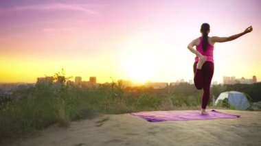 Genç bir kadın büyük bir şehir arka planda bir dağda yoga uygulamaları. Sağlıklı kadın günbatımında spor yapıyor. Kadın atlet pembe gömlek ve mor tayt güneş doğarken bir ısınma yok.