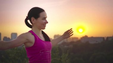 Genç bir kadın büyük bir şehir arka planda bir dağda yoga uygulamaları. Sağlıklı kadın günbatımında spor yapıyor. Kadın atlet pembe gömlek ve mor tayt güneş doğarken bir ısınma yok.