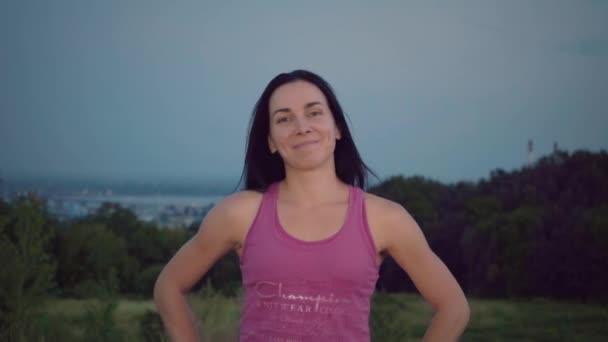 Portret atrakcyjna młoda kobieta na tle miasta. Sportsmenki w fioletowy t-shirt pokazuje ramionach athletic, uśmiechając się do kamery. Ładna, młoda kobieta z sportowy kompilacji. Długowłosy Brunetki — Wideo stockowe