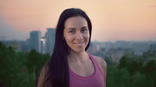 Retrato de una atractiva deportista en el fondo de la ciudad. Linda mujer joven con una construcción deportiva está mirando a la cámara. Morena de pelo largo posando sobre el telón de fondo de la puesta del sol — Vídeos de Stock