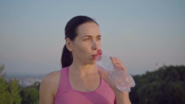 Porträt eines attraktiven Mädchens vor dem Hintergrund der Stadt. Sportlerin im lila T-Shirt trinkt Wasser aus einer Flasche. Die hübsche junge Frau mit sportlicher Statur blickt in die Kamera. langhaarig — Stockvideo