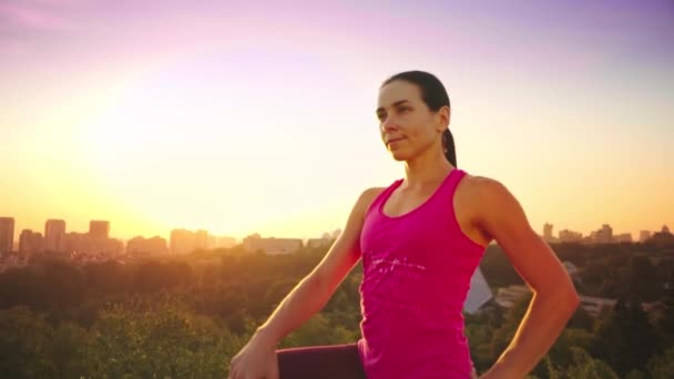 Egy fiatal nő egy hegy, a háttérben egy nagy város jógázik. Egészséges nő sportot a naplemente. A női sportoló, rózsaszín inget és lila leggings csinál bemelegítéssel napkeltekor. — Stock videók