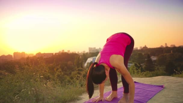 En ung kvinne øver på yoga på et fjell i bakgrunnen av en storby. Frisk kvinne driver med sport ved solnedgang. En kvinnelig idrettsutøver i rosa skjorte og lilla bukser varmer opp ved soloppgang . – stockvideo