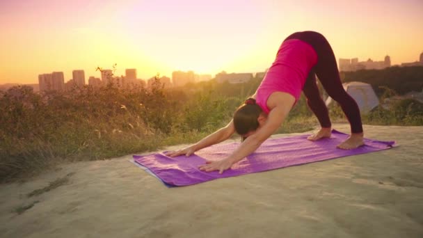 Genç bir kadın büyük bir şehir arka planda bir dağda yoga uygulamaları. Sağlıklı kadın günbatımında spor yapıyor. Kadın atlet pembe gömlek ve mor tayt güneş doğarken bir ısınma yok. — Stok video