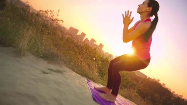 A young woman practices yoga on a mountain in the background of a big city. Healthy woman doing sports at sunset. A female athlete in a pink shirt and purple leggings does a warm-up at sunrise. — Stock Video