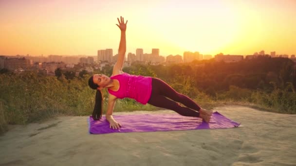 Genç bir kadın büyük bir şehir arka planda bir dağda yoga uygulamaları. Sağlıklı kadın günbatımında spor yapıyor. Kadın atlet pembe gömlek ve mor tayt güneş doğarken bir ısınma yok. — Stok video