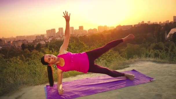 Genç bir kadın büyük bir şehir arka planda bir dağda yoga uygulamaları. Sağlıklı kadın günbatımında spor yapıyor. Kadın atlet pembe gömlek ve mor tayt güneş doğarken bir ısınma yok. — Stok video