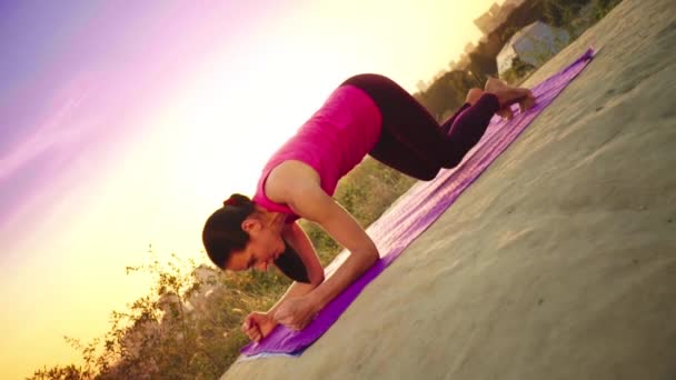 Una joven practica yoga en una montaña en el fondo de una gran ciudad. Mujer sana haciendo deportes al atardecer. Una atleta con camisa rosa y polainas moradas se calienta al amanecer . — Vídeo de stock
