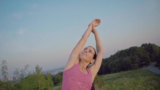 En ung kvinna övar yoga på ett berg i bakgrunden av en storstad. Frisk kvinna gör sport vid solnedgången. En kvinnlig idrottare i en rosa skjorta och lila leggings gör en uppvärmning på sunrise. — Stockvideo