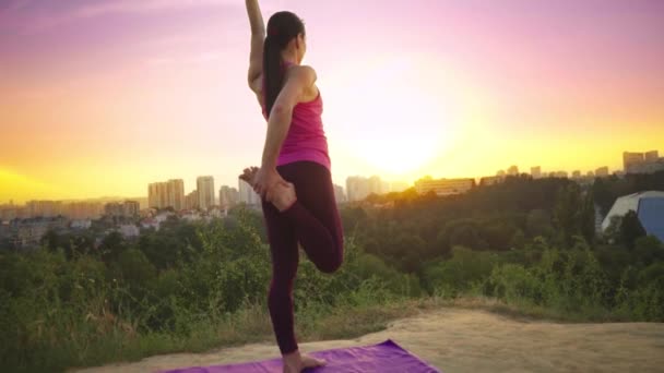 Een jonge vrouw beoefent yoga op een berg op de achtergrond van een grote stad. Gezonde vrouw sport doen bij zonsondergang. Een vrouwelijke atleet in een roze shirt en Paarse legging is een warming-up bij zonsopgang. — Stockvideo