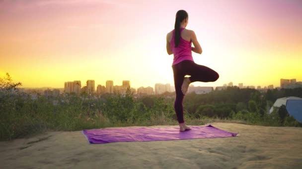 En ung kvinna övar yoga på ett berg i bakgrunden av en storstad. Frisk kvinna gör sport vid solnedgången. En kvinnlig idrottare i en rosa skjorta och lila leggings gör en uppvärmning på sunrise. — Stockvideo