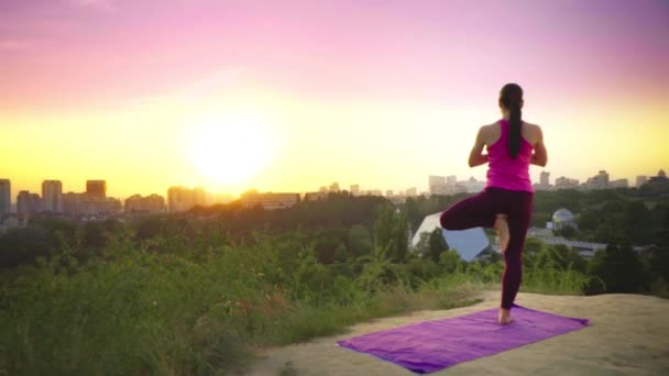 Genç bir kadın büyük bir şehir arka planda bir dağda yoga uygulamaları. Sağlıklı kadın günbatımında spor yapıyor. Kadın atlet pembe gömlek ve mor tayt güneş doğarken bir ısınma yok. — Stok video