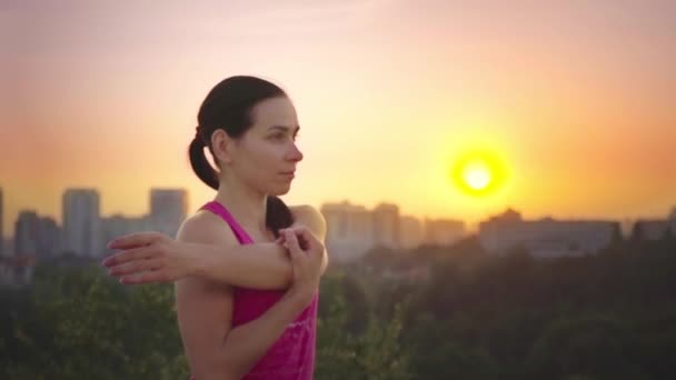 Een jonge vrouw beoefent yoga op een berg op de achtergrond van een grote stad. Gezonde vrouw sport doen bij zonsondergang. Een vrouwelijke atleet in een roze shirt en Paarse legging is een warming-up bij zonsopgang. — Stockvideo