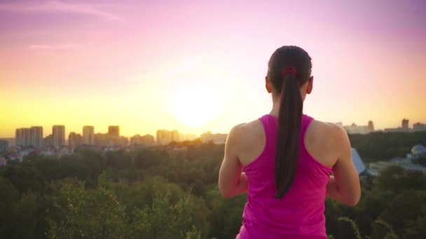 Een jonge vrouw beoefent yoga op een berg op de achtergrond van een grote stad. Gezonde vrouw sport doen bij zonsondergang. Een vrouwelijke atleet in een roze shirt en Paarse legging is een warming-up bij zonsopgang. — Stockvideo