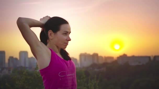 En ung kvinna övar yoga på ett berg i bakgrunden av en storstad. Frisk kvinna gör sport vid solnedgången. En kvinnlig idrottare i en rosa skjorta och lila leggings gör en uppvärmning på sunrise. — Stockvideo