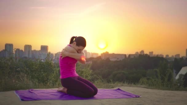 En ung kvinne øver på yoga på et fjell i bakgrunnen av en storby. Frisk kvinne driver med sport ved solnedgang. En kvinnelig idrettsutøver i rosa skjorte og lilla bukser varmer opp ved soloppgang . – stockvideo