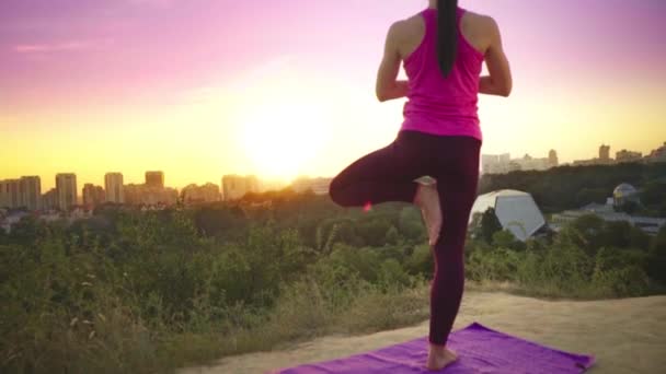 En ung kvinna övar yoga på ett berg i bakgrunden av en storstad. Frisk kvinna gör sport vid solnedgången. En kvinnlig idrottare i en rosa skjorta och lila leggings gör en uppvärmning på sunrise. — Stockvideo