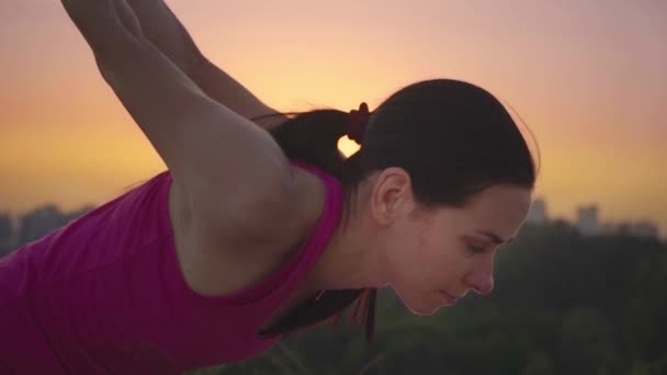 Una joven practica yoga en una montaña en el fondo de una gran ciudad. Mujer sana haciendo deportes al atardecer. Una atleta con camisa rosa y polainas moradas se calienta al amanecer . — Vídeos de Stock