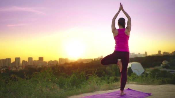 Una giovane donna pratica yoga su una montagna sullo sfondo di una grande città. Donna sana che fa sport al tramonto. Un'atleta donna in camicia rosa e leggings viola fa un riscaldamento all'alba . — Video Stock