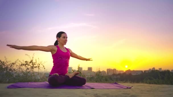 Una joven practica yoga en una montaña en el fondo de una gran ciudad. Mujer sana haciendo deportes al atardecer. Una atleta con camisa rosa y polainas moradas se calienta al amanecer . — Vídeos de Stock