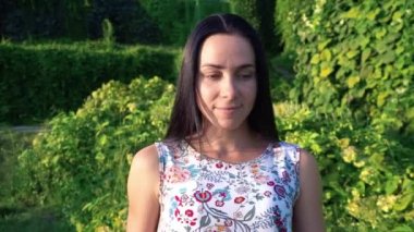 Yeşil park karşı çukurlu yanakları güzel bir genç kadın portresi. Yavaş yavaş esmer gülümseyen bahçede yeşil çalılar boyunca yol gösterir. Çiçek baskı ile yaz elbiseli güzel kız