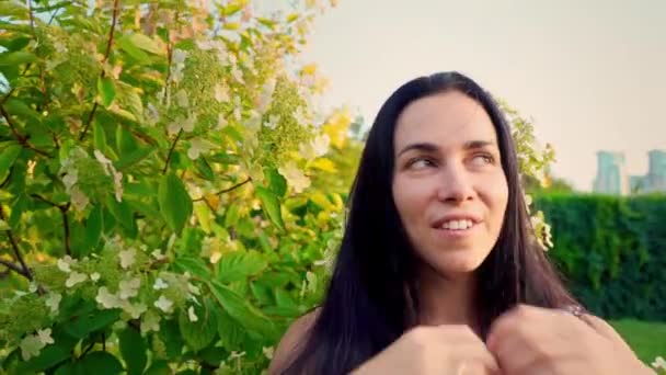 Portret van een lachende brunette met golvend wangen tegen een groen park. Meisje kijkt naar de camera en spelen met haar haren. Mooie jonge vrouw staat in park en kijkt naar de camera — Stockvideo