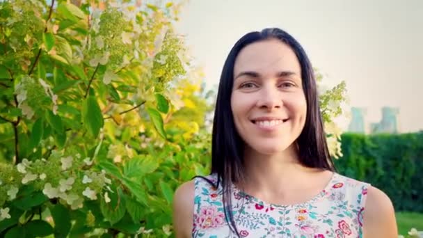 Ritratto di una bruna sorridente dalle guance rammollite contro un parco verde. Bella giovane donna si trova nel parco e guarda la fotocamera. Bella ragazza in abito estivo con stampa floreale — Video Stock