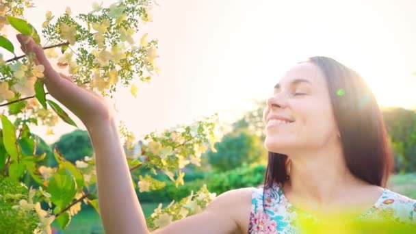 Retrato de uma morena sorridente com bochechas onduladas contra um parque verde. Mulher bonita fica no parque e olha para a câmera. Menina bonita em vestido de verão com estampa floral inala o cheiro de um — Vídeo de Stock