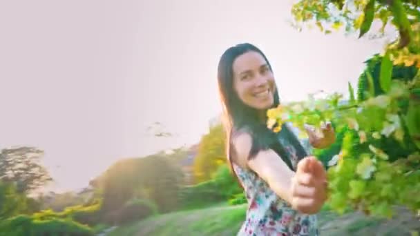 Jovem mulher brinca corre em torno de uma árvore verde no jardim. Retrato de uma morena sorridente com bochechas onduladas contra um parque verde. Mulher bonita olha para a câmera. Menina bonita no verão — Vídeo de Stock