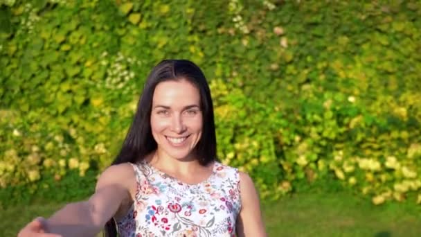European young woman with long black hair runs away from the camera in slow motion, calling for herself. Attractive girl with dimpled cheeks in a white floral print beckons to follow her. Smiling — Stock Video