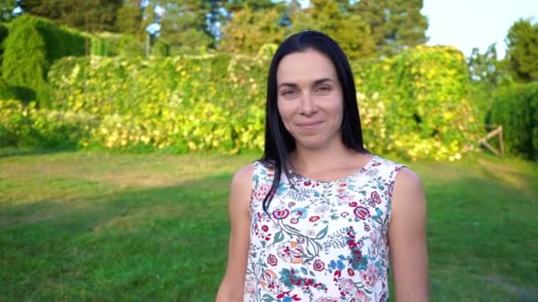 Europese jonge vrouw met lange zwarte haren loopt weg van de camera in slow motion, bellen voor zichzelf. Aantrekkelijk meisje met golvend wangen in een witte bloemen print wenkt te volgen haar. Glimlachend — Stockvideo