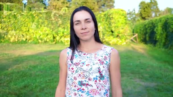 Ritratto di una bruna sorridente dalle guance rammollite contro un parco verde. Bella giovane donna si trova nel parco e guarda la fotocamera. Bella ragazza in abito estivo con stampa floreale — Video Stock