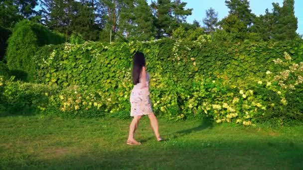 Jonge vrouw speels cirkelen rond zichzelf in een groene tuin. Portret van een lachende brunette met golvend wangen tegen een groen park. Mooie jonge vrouw wordt gekeken naar de camera. Mooi meisje in de zomer — Stockvideo