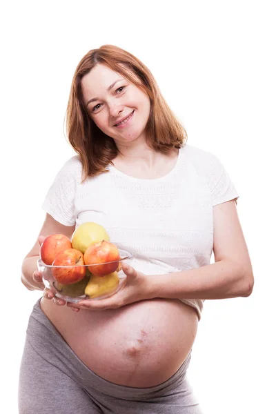 Mulher grávida sorrindo para a câmera e segurando uma tigela de vidro com frutas em suas mãos — Fotografia de Stock