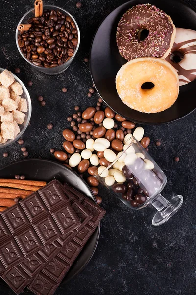 Κάτοψη των φυστικιών σοκολάτας, χύθηκε στο σκούρο ταμπλό, δίπλα σε δισκία σοκολάτας, ντόνατς, καφέ ζάχαρη και κόκκους καφέ — Φωτογραφία Αρχείου